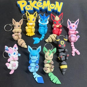 Könnte beinhalten: Eine Sammlung farbenfroher Pokémon-Charakter-Schlüsselanhänger. Die Schlüsselanhänger zeigen verschiedene Pokémon-Designs in leuchtenden Farben, darunter Gelb, Blau, Rot, Rosa und Grün. Das Wort "Pokémon" wird in blauen und gelben Buchstaben über den Schlüsselanhängern angezeigt.