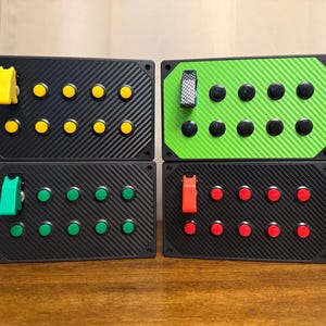 May include: Four rectangular button boxes with carbon fiber textured faces. Each box features a single toggle switch and ten circular push buttons. The top left box is black with yellow buttons, top right is green with black buttons, bottom left is black with green buttons, and bottom right is black with red buttons.