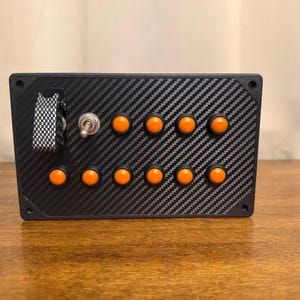 May include: A rectangular control panel featuring a black carbon fiber pattern finish. The device includes ten round orange push buttons arranged in two rows, a single metal toggle switch, and a black and white checkered safety flip cover.