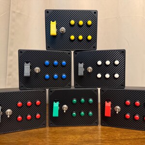 May include: Six rectangular black control boxes with a carbon fiber texture, each featuring a central metal toggle switch, a colored safety flip cover, and six small round push buttons in matching colors including yellow, blue, white, red, and green.