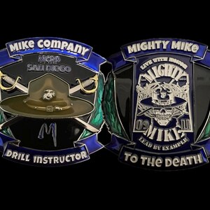 May include: A black and green military challenge coin with a blue ribbon. The front side features a green military hat with crossed swords and the text "Mike Company MCRD San Diego Drill Instructor". The back side features a skull with crossed swords and the text "Mighty Mike Live With Honor 09 Mighty III Lead By Example To The Death".