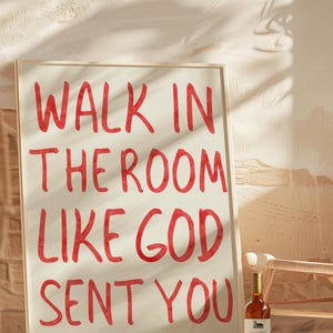 Puede incluir: Un cartel enmarcado con el texto "WALK IN THE ROOM LIKE GOD SENT YOU" en letras rojas. Una botella de vino está junto al cartel. El fondo es una pared texturizada con sombras.