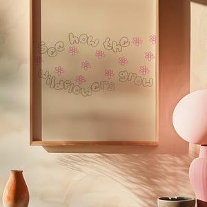 May include: A framed print with the words "See how the wildflowers grow" in a whimsical font, accompanied by pink flower illustrations. The frame is made of light-colored wood. The print is displayed on a pink wall, with decorative vases and a bowl in the foreground.