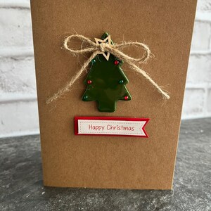 May include: A brown card with a green Christmas tree made of ceramic with a wooden star on top. The tree is decorated with red and green beads. The card has a red banner with the text "Happy Christmas" on it.