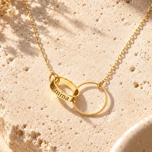 May include: A gold-toned necklace featuring a delicate chain connected to two interlocking circular pendants. One pendant is a smooth, thin ring, while the other is a slightly thicker, flattened oval band engraved with the name Emma in black text.
