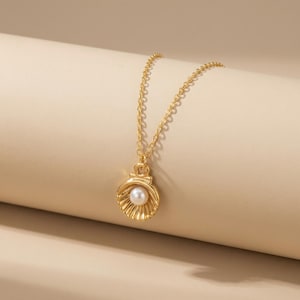 Könnte beinhalten: Eine goldfarbene Halskette mit einem Muschel-Anhänger und einer Perle. Der Anhänger hat ein detailliertes, strukturiertes Design. Die Kette ist filigran und ebenfalls goldfarben. Die Halskette wird vor einem neutralen Hintergrund gezeigt.