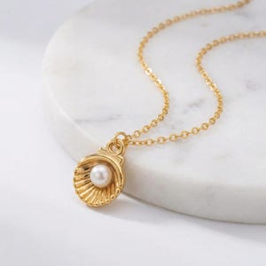 May include: A gold-colored necklace featuring a seashell pendant with a pearl center. The pendant is detailed with ribbed lines, and the chain is delicate. The necklace rests on a white marble surface.