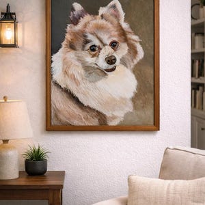 May include: A framed portrait of a fluffy, light brown and white dog. The dog has a friendly expression and is set against a dark background. The artwork is displayed on a white wall above a beige sofa.
