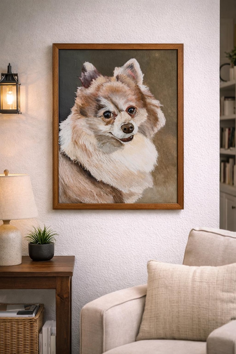 May include: A framed portrait of a fluffy, light brown and white dog. The dog has a friendly expression and is set against a dark background. The artwork is displayed on a white wall above a beige sofa.