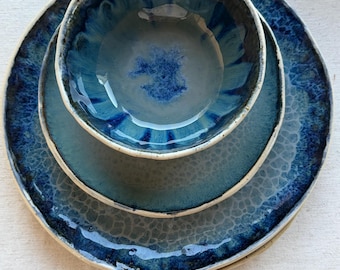 May include: A ceramic dinnerware set featuring a large plate, a smaller plate, and a bowl. The set has a glossy, reactive glaze in shades of blue and teal, with a textured, crackle-like pattern. The rims are a lighter color.