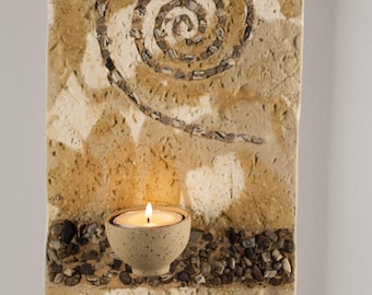 May include: A rectangular, textured wall-mounted candle holder. The holder features a spiral design made of small stones and a speckled ceramic bowl holding a lit candle. The color palette includes beige, brown, and gray.