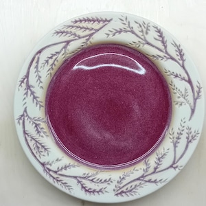 以下が含まれることがあります： 深いバーガンディ色の中心部と白い縁を持つセラミックプレート。縁は紫色の葉の繰り返し模様で装飾されています。陶器の道具が背景に置かれています。