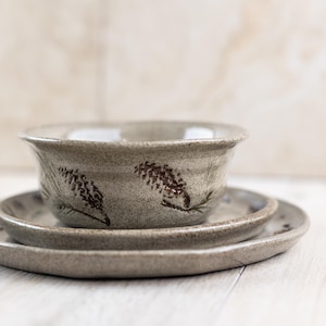 Puede incluir: Un juego de tres platos de cerámica de color gris claro con un diseño floral marrón. Los platos están anidados, con el plato más pequeño en la parte superior.