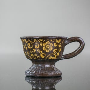 May include: A brown ceramic teacup with a gold floral design. The cup has a pedestal base and a handle.