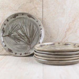 May include: A set of ceramic plates featuring a pine needle design. The plates are a light grey colour with brown accents. The plates are stacked on top of each other.