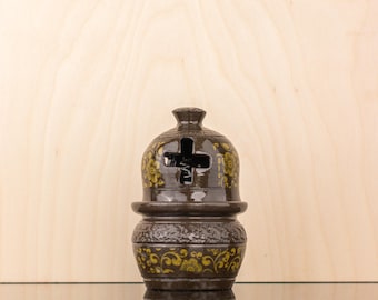 May include: A brown ceramic incense burner with a cross-shaped opening and gold floral designs. The burner is sitting on a reflective surface.