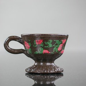 May include: A brown ceramic teacup with a floral design featuring red roses and green leaves. The cup has a handle and a pedestal base.