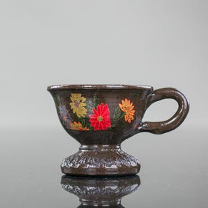 May include: A small brown ceramic cup with a floral design. The cup has a wide base and a handle. The flowers are painted in red, yellow, and green.