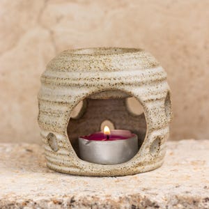 Puede incluir: Un difusor de aceite de cerámica blanca con forma redonda y superficie texturizada. El difusor tiene una pequeña abertura en el centro para una vela de té. Una vela de té rosa se coloca dentro del difusor.