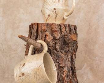 May include: Two beige ceramic mugs with botanical designs. One mug rests on a rustic tree stump, while the other hangs from a branch attached to the stump. The mugs have a speckled texture and curved handles.