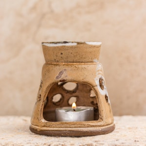 Puede incluir: Un difusor de aceite de cerámica marrón con un glaseado blanco y una vela pequeña en el interior. El difusor tiene una abertura circular en el centro y un patrón decorativo de agujeros alrededor de la abertura.