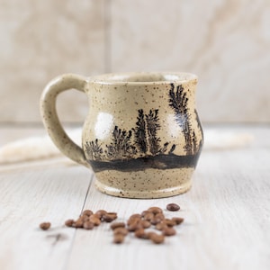 May include: A beige ceramic mug with a black and white tree design. The mug has a speckled glaze and a rounded body. The mug is sitting on a light wood surface with coffee beans scattered around it.