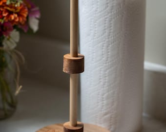 May include: A wooden paper towel holder with a round base and a vertical dowel. The holder has a natural wood finish and is designed to hold a standard roll of paper towels. A roll of white paper towels is next to the holder.