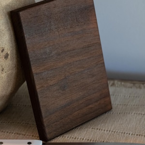 May include: A rectangular, dark brown wooden cutting board with visible wood grain. A white-handled knife with a silver blade rests on a beige surface in front of the board. The board is leaning against a beige vase.