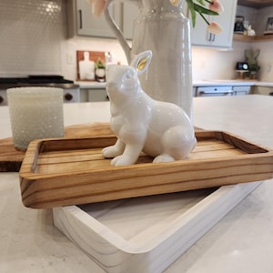 May include: A white ceramic rabbit figurine with gold accents sits on a wooden tray with a white tray underneath. The tray is on a white countertop.