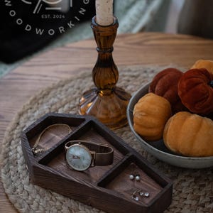 May include: A wooden coffin-shaped trinket tray with compartments for jewelry. The tray is made of dark wood and has a natural finish. It is sitting on a woven rug on a wooden table. The tray is filled with a watch, a bracelet, and earrings.