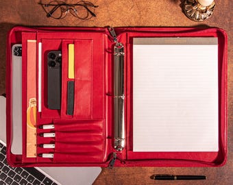 May include: A red leather portfolio with a zipper closure, open to reveal a notepad, phone, tablet, pens, and various pockets. A pair of round eyeglasses and a laptop are visible. The portfolio is on a brown surface.