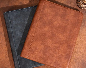 May include: Two leather document holders. One is a dark gray-blue color with light blue stitching, and the other is a warm brown with matching stitching. The folders are rectangular and appear to be closed.