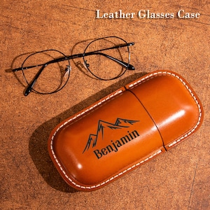 Puede incluir: Un estuche de cuero color canela para gafas con costuras blancas, que presenta un gráfico de montaña negro y el nombre Benjamin impreso en el frente. Un par de gafas de metal negro con montura fina descansa arriba. El texto Leather Glasses Case es visible.