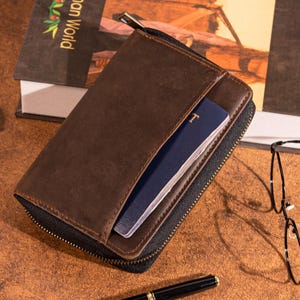 May include: A brown leather wallet with a black zipper, open to reveal a blue passport. The wallet is rectangular and appears to be made of high-quality leather. A pen and glasses are in the background.
