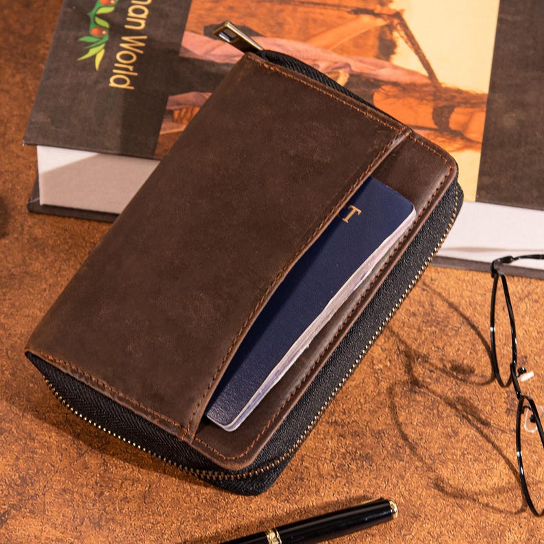 May include: A brown leather wallet with a black zipper, open to reveal a blue passport. The wallet is rectangular and appears to be made of high-quality leather. A pen and glasses are in the background.