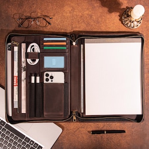 May include: Open brown leather portfolio with multiple compartments for a tablet, cards, phone, pens, and a notepad. The portfolio is open, revealing the interior with various items, including a white phone and a notepad with grid paper.