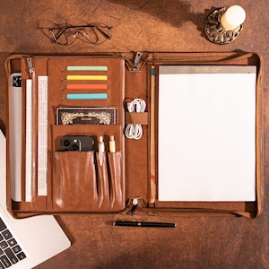 May include: A brown leather portfolio with multiple compartments, including card slots, pen holders, and a notepad. The portfolio is open, revealing a tablet, phone, pens, and a black pen. A pair of glasses and a candle holder are also visible.