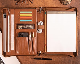 May include: A brown leather portfolio with multiple compartments, including card slots, pen holders, and a notepad. The portfolio is open, revealing a tablet, phone, pens, and a black pen. A pair of glasses and a candle holder are also visible.
