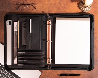 May include: A black leather binder with a copper-colored spine and zipper, open to reveal a notepad and interior pockets. A laptop, glasses, and a pen are also on the wooden surface. The binder contains the text "Petra Diamonds".