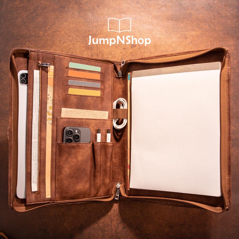 May include: A brown leather zippered portfolio with multiple pockets and slots for holding a tablet, phone, pens, and other accessories. The portfolio is open to reveal a white notepad with a grid pattern.