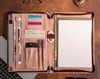 May include: A pink leather portfolio with a zipper closure, open to reveal a notepad, phone, pens, and card slots. A laptop and glasses are also visible. The portfolio is on a brown surface.