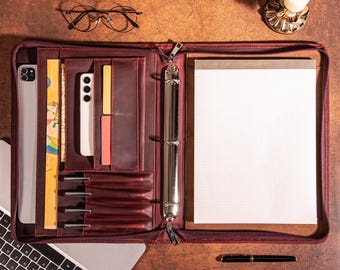 May include: A burgundy leather portfolio with a zipper closure, open to reveal a notepad, pen, and various pockets holding a tablet, phone, and cards. A pair of glasses and a candle holder are on the table.
