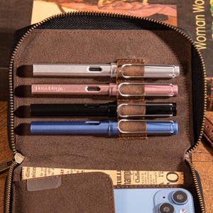 May include: Four fountain pens in silver, rose gold, black, and blue are displayed in a brown leather pen case. Each pen has a silver nib and a brown leather pen clip. The case is open, revealing the pens and a small pocket.