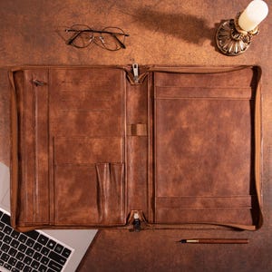 May include: A brown leather portfolio with multiple pockets and a zipper closure. The portfolio is open and lying flat on a brown surface.