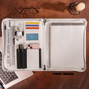 May include: A white zippered portfolio organizer featuring multiple interior slots for credit cards, a clear ID window, a dedicated pocket for a smartphone, two pen loops, a white charging cable, and a lined notepad on the right side.