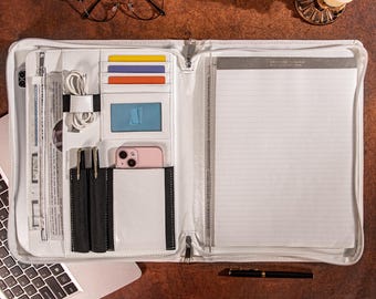 May include: A white zippered portfolio organizer featuring multiple interior slots for credit cards, a clear ID window, a dedicated pocket for a smartphone, two pen loops, a white charging cable, and a lined notepad on the right side.