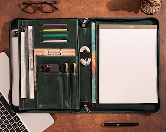 May include: A dark green leather portfolio with multiple compartments, including card slots, pen holders, and a notepad. The portfolio is open, revealing a smartphone, pens, and a tablet. A laptop and glasses are also visible.
