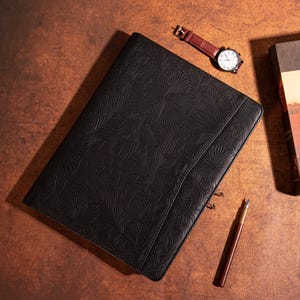 Op de afbeelding: Zwarte lederen map met een reliëf van bloemen. De map heeft een metalen sluiting en een pennenlus.