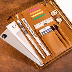 May include: A brown leather portfolio with multiple compartments. It contains a tablet, a smartphone, pens, and a newspaper clipping with the text "making us all fat?". There are also colorful sticky notes and a charging cable.