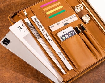 May include: A brown leather portfolio with multiple compartments. It contains a tablet, a smartphone, pens, and a newspaper clipping with the text "making us all fat?". There are also colorful sticky notes and a charging cable.